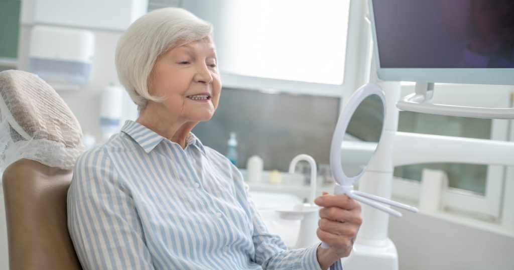 The senior patient is looking at her teeth in the mirror in a dental office, representing CDCP dental implant coverage